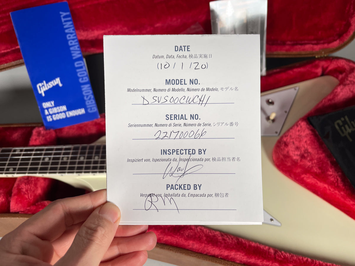 A hand holds a white inspection card over a white guitar body in a red plush case, with a blue warranty card visible.
