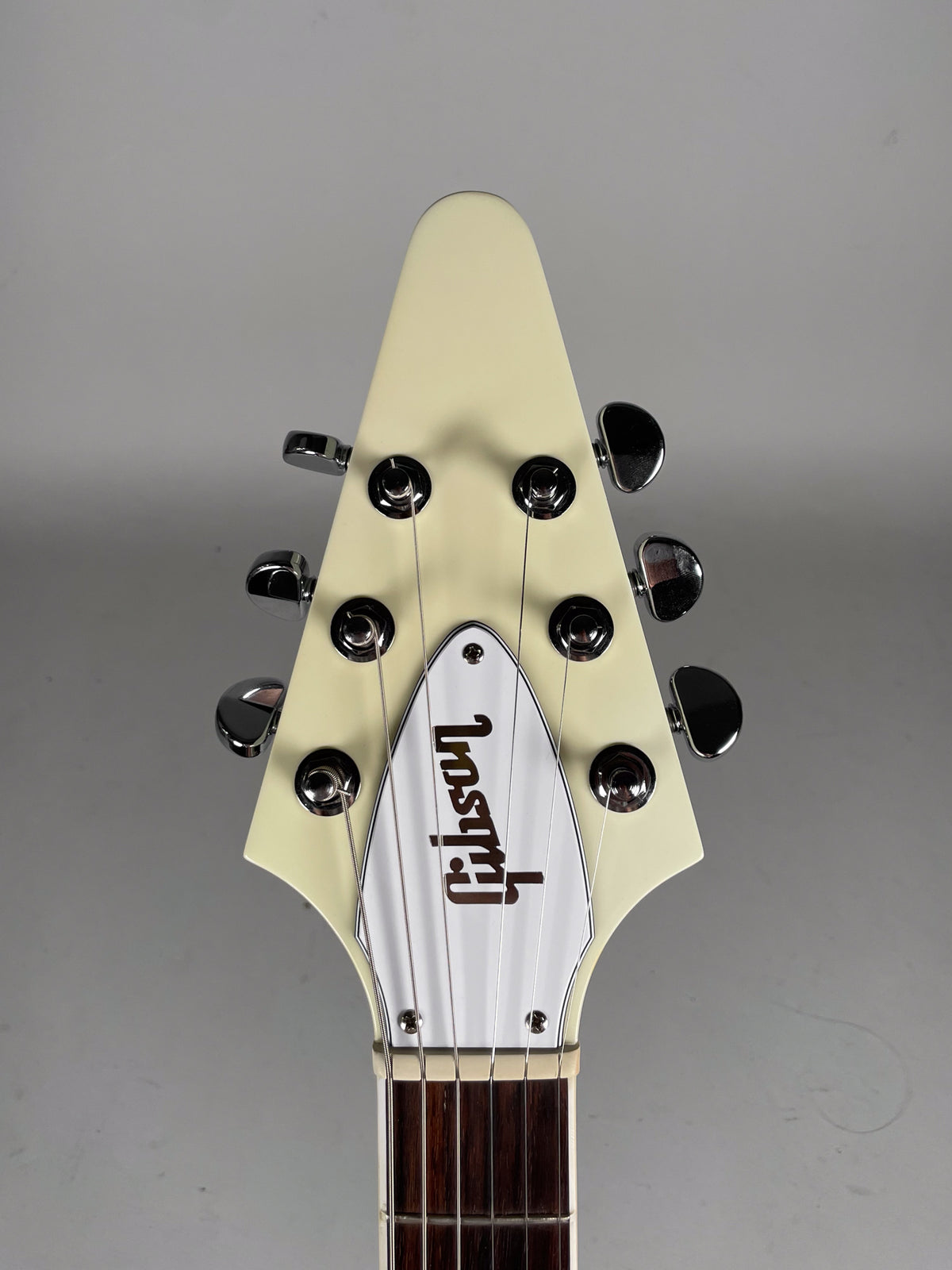 The Flying V's cream-white headstock, with six chrome tuners and a white truss rod cover, is centered against a grey background.