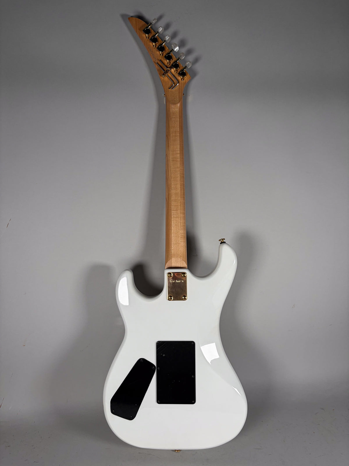 The back of a white electric guitar with a light wood neck, gold neck plate, and two black control cavity covers.
