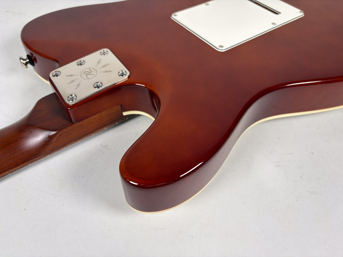 Close-up of the back of a reddish-brown guitar, showing the neck joint, a chrome neck plate with a logo, and a white control cavity cover.