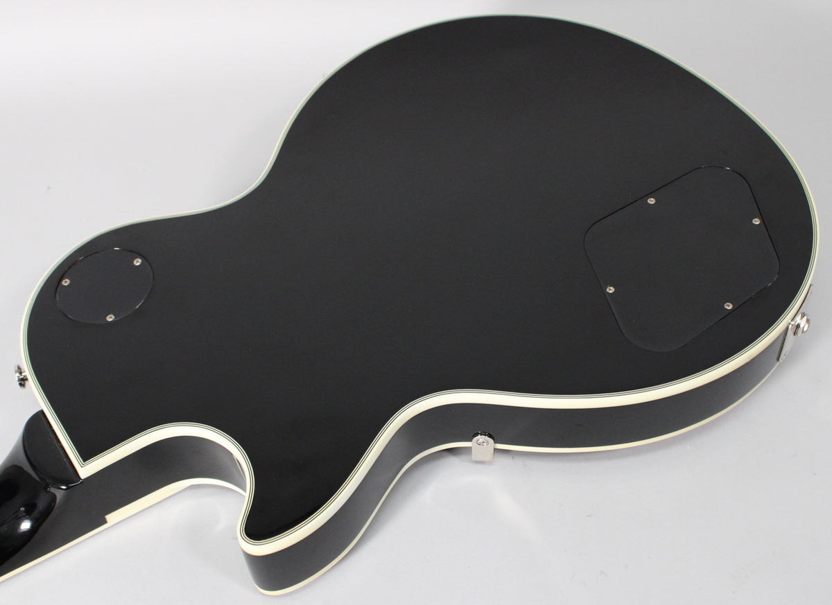 The guitar's matte black back is angled, showing cream binding, a round control cavity cover, and a rectangular electronics cover.