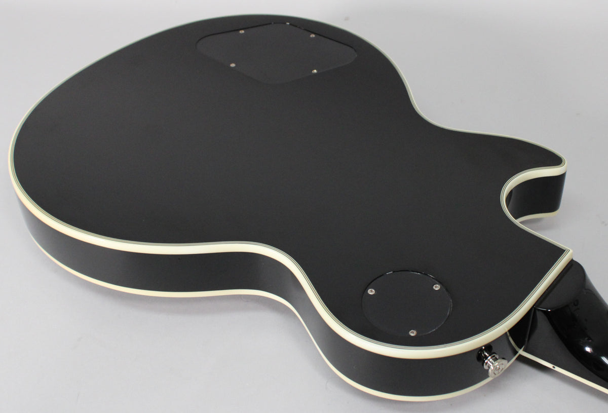 The guitar's matte black back is angled, showing cream binding, two control cavity covers, and a chrome strap button.