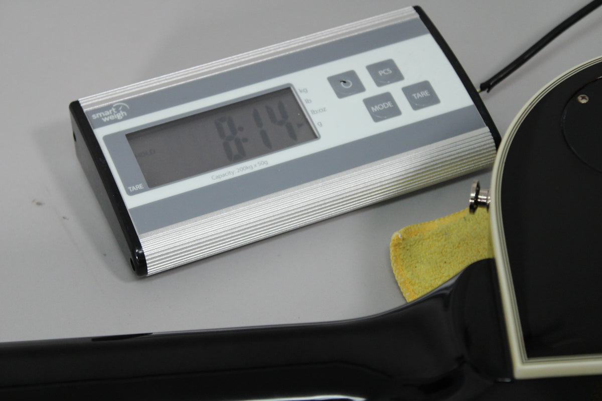 The guitar's black back and cream binding rest on a scale displaying "8:14", with a yellow cloth under the strap button.