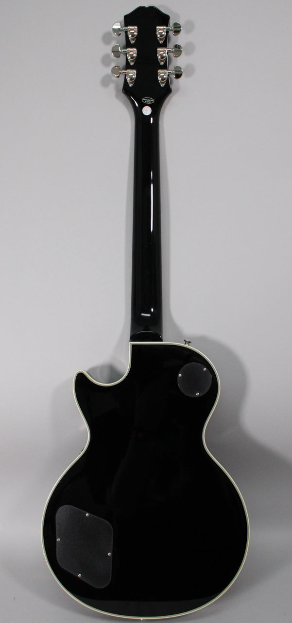 Full rear view of a black gloss Les Paul, showing the headstock with six chrome tuners, neck, and body with two control cavity covers.