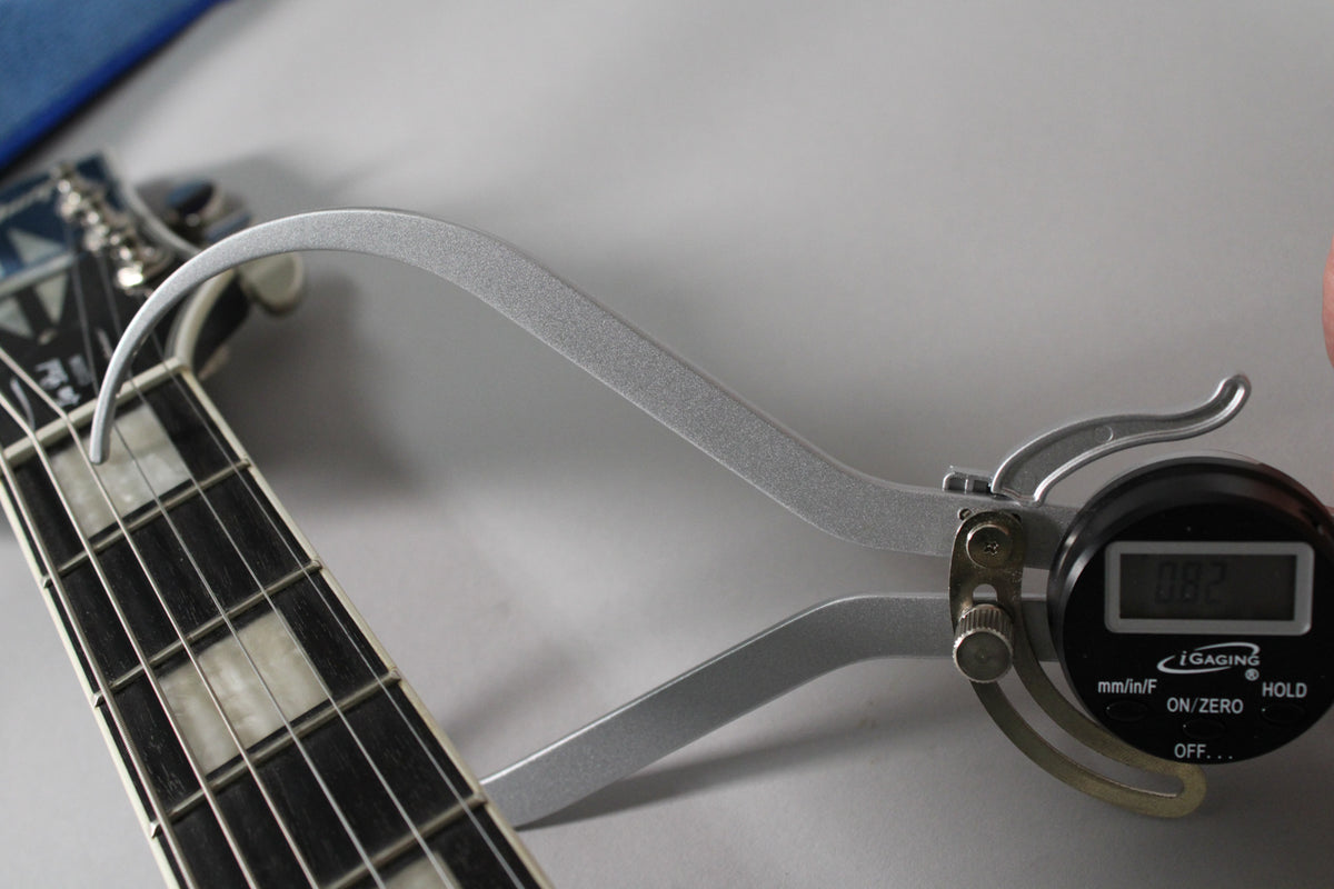 Close-up of a digital caliper measuring the nut height on the guitar's dark fretboard with rectangular pearloid inlays.