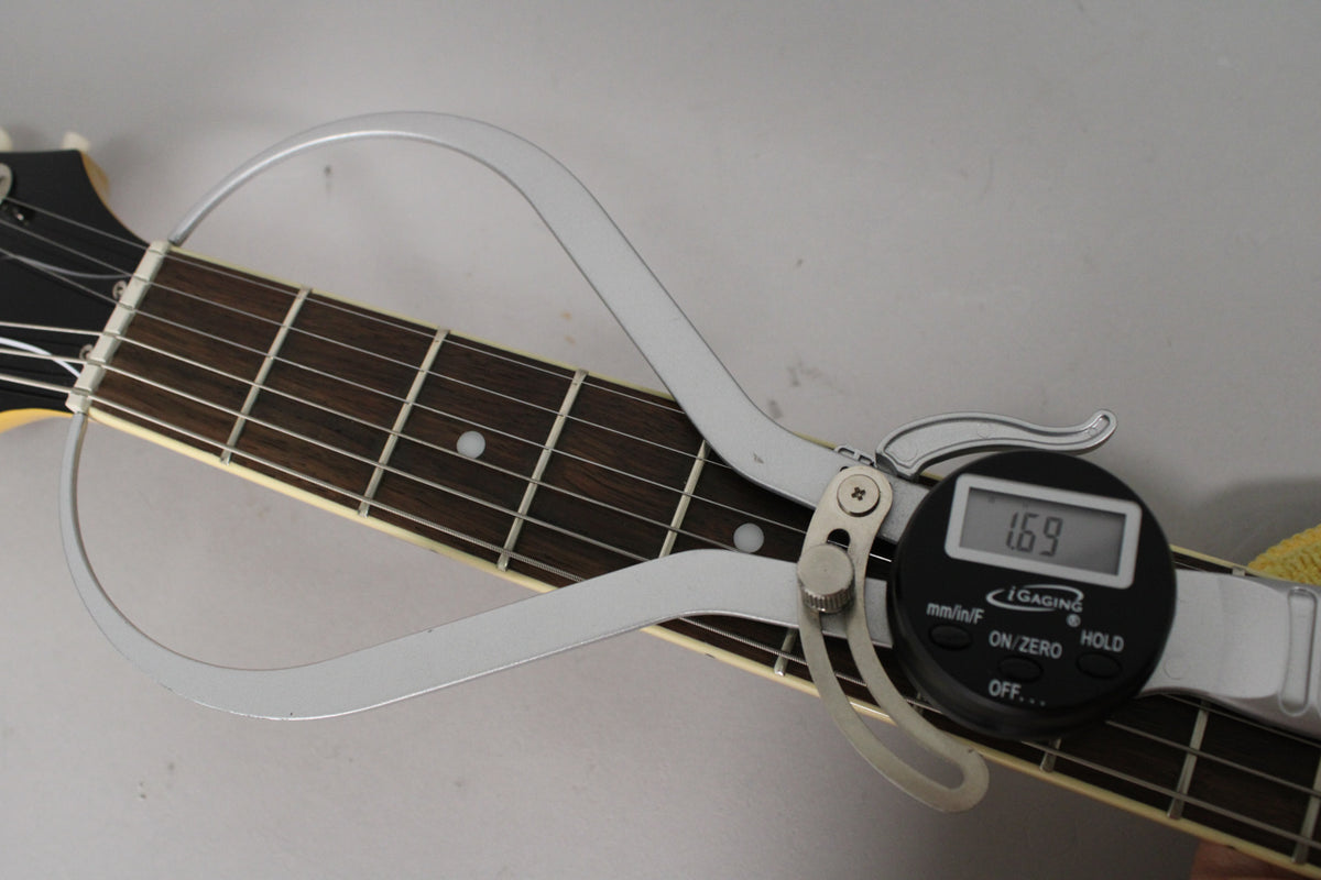 A digital caliper measures the nut width of the guitar neck, displaying "1.69" against the dark fretboard and light binding.