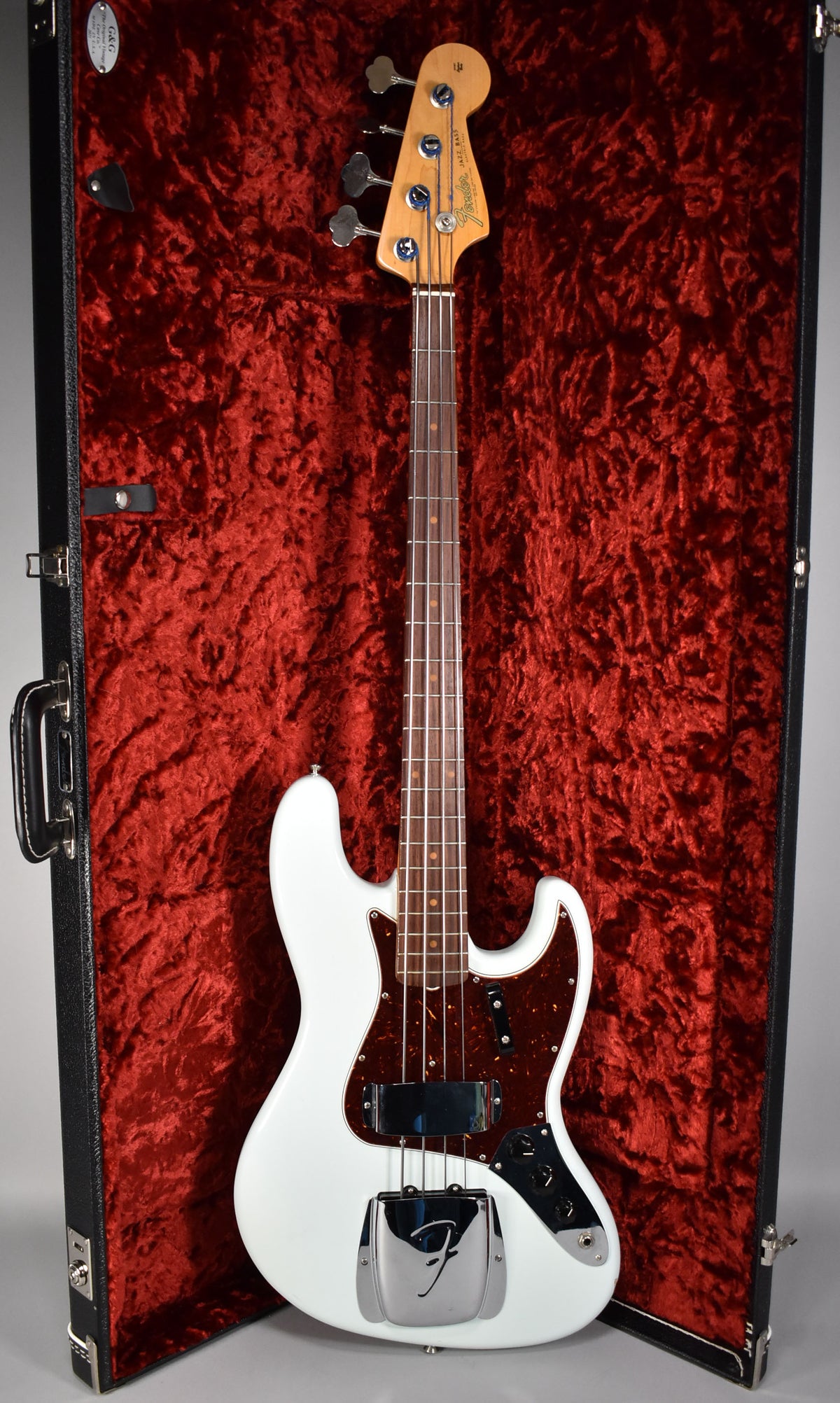 A white Fender Jazz Bass with a tortoiseshell pickguard and chrome hardware, resting in a black hard case with red velvet lining.