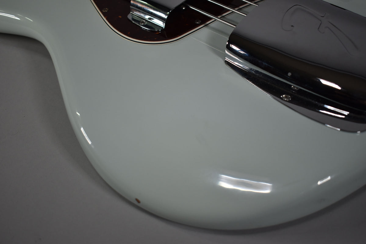 Close-up of the bass's glossy sonic blue body, showing the chrome bridge cover, brown pickguard, and a small red mark on the lower bout.