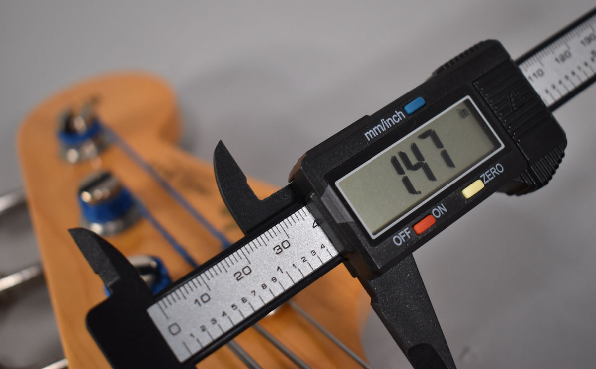 A digital caliper measures the bass's nut width at 1.47 inches, with the headstock and tuning pegs blurred in the background.