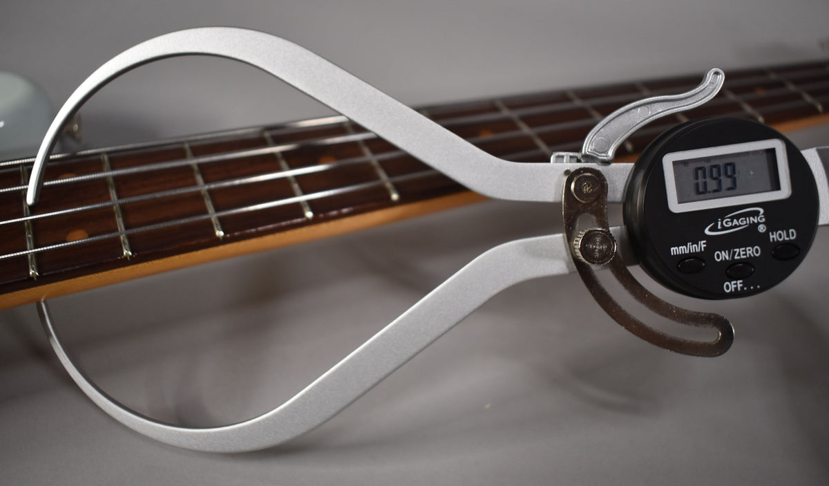 A digital caliper measures the string height at the 12th fret of the bass, displaying "0.99" on its screen.