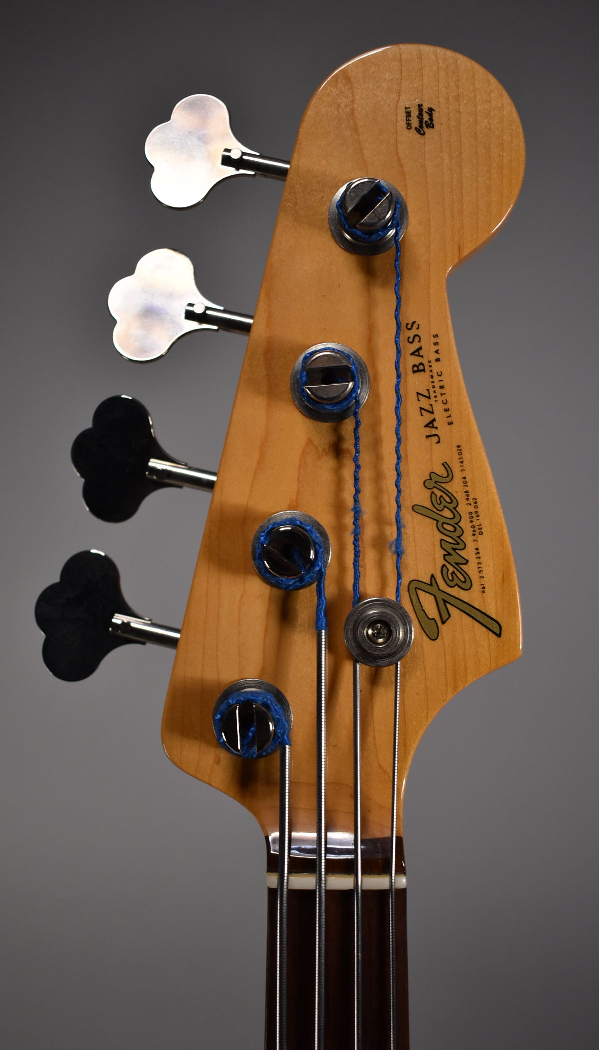 Close-up of the bass headstock, showing the light wood finish, four metal tuners, and blue string windings.