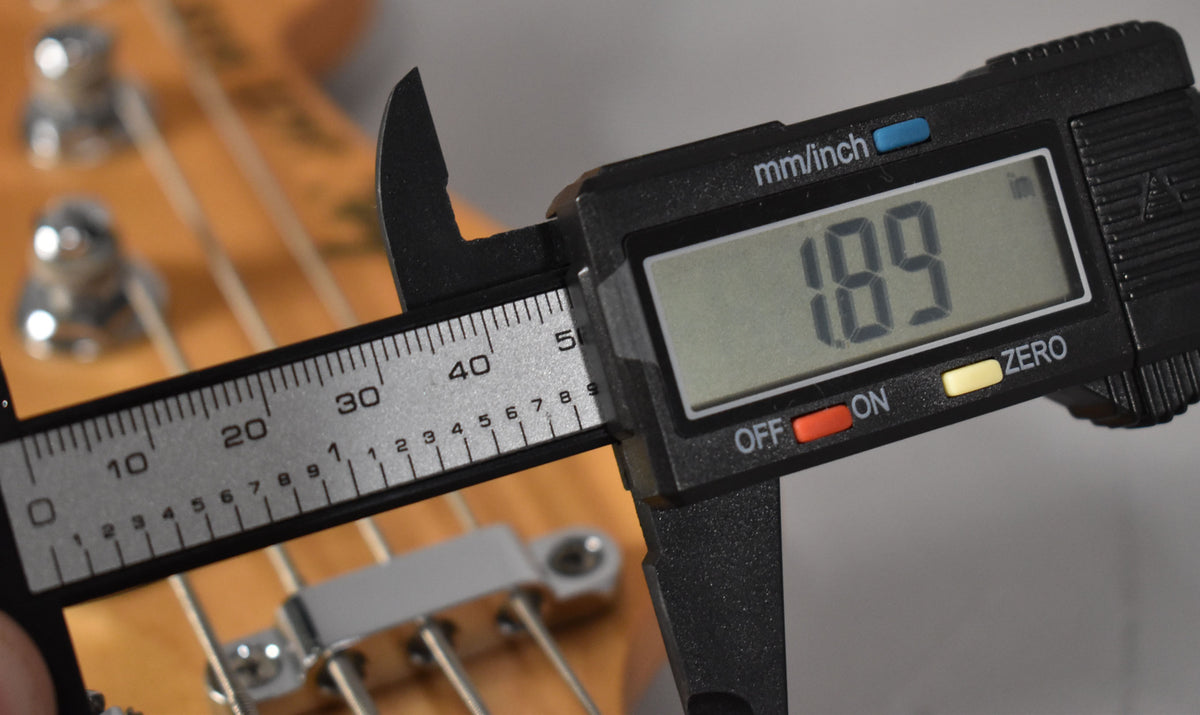 A digital caliper measures the nut width of the bass, showing 1.89 inches, with the light maple neck and strings visible behind it.