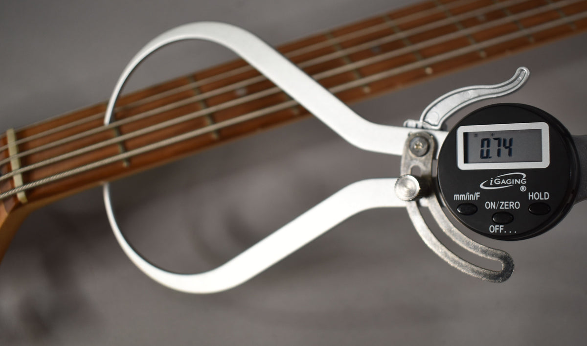 A digital caliper measures the bass's fretboard, showing "0.74" on its screen, with five strings visible.