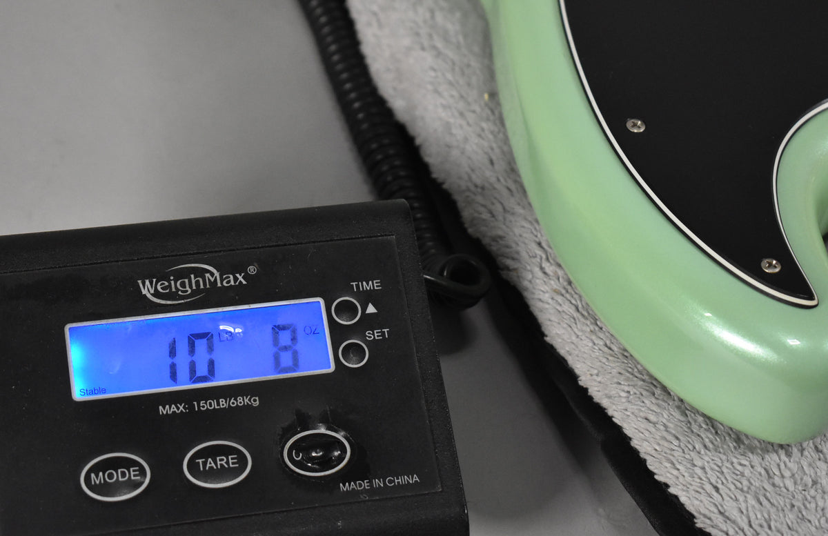 Close-up of a black digital scale displaying "10 LBS 8 OZ" next to the shiny Surf Pearl green bass body and black pickguard.