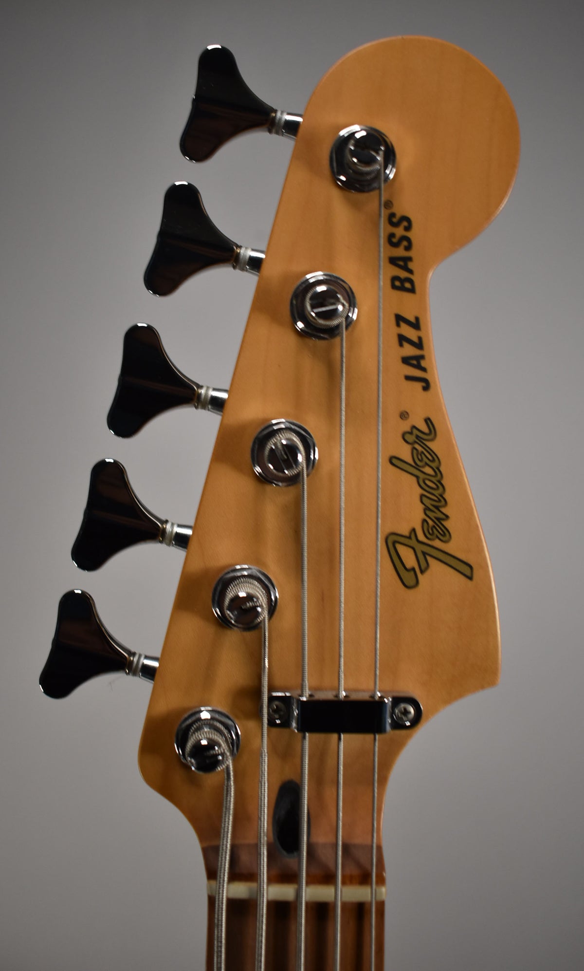 Close-up of the bass headstock, angled slightly left, showing five chrome tuners on a light wood finish, with no visible wear.