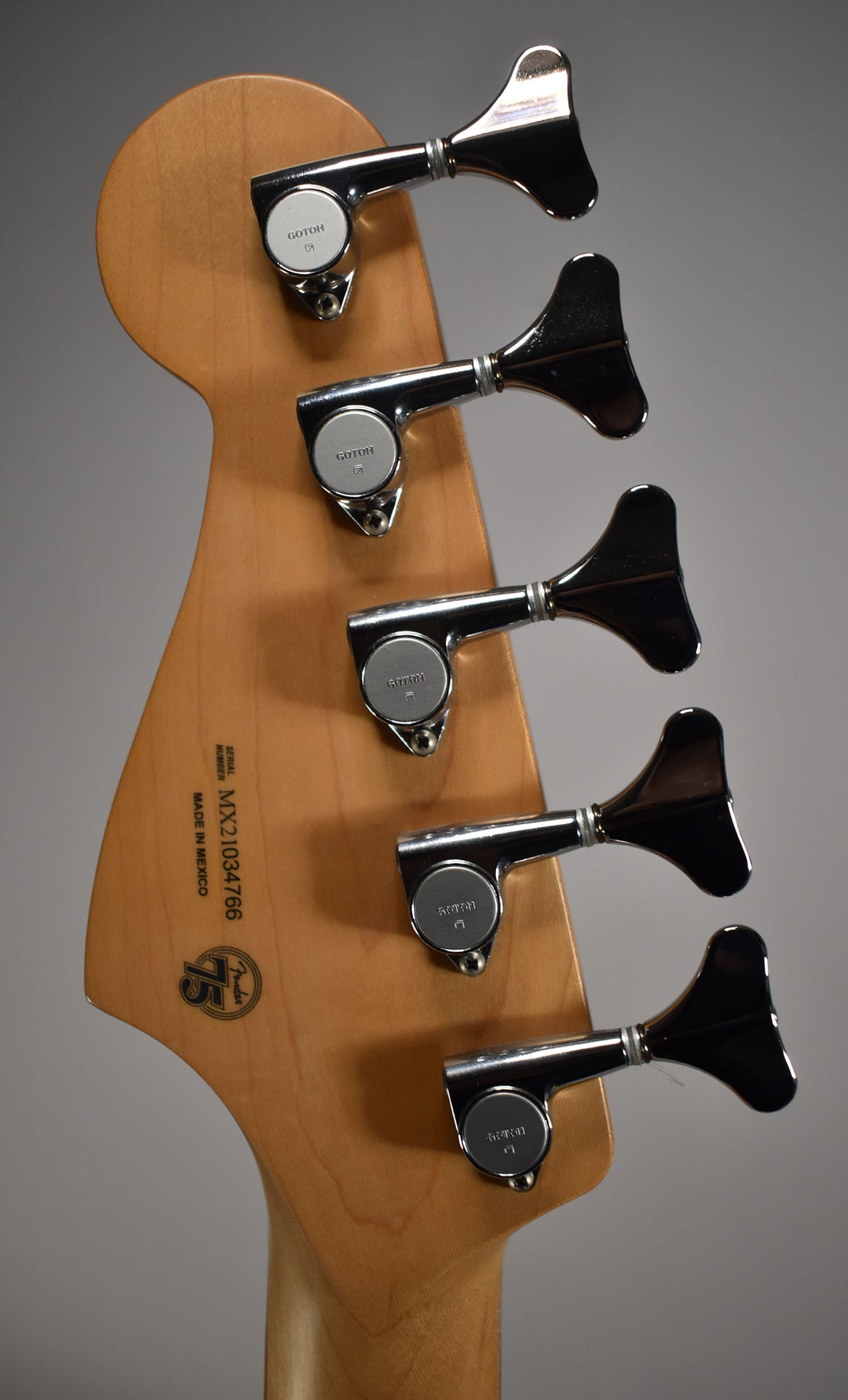 The back of a light wood bass headstock with five chrome tuners and a "75" anniversary logo.