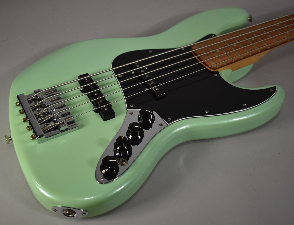 Close-up of a mint green bass guitar, angled from the lower bout, showing the chrome bridge, black pickups, and control plate with five black knobs.