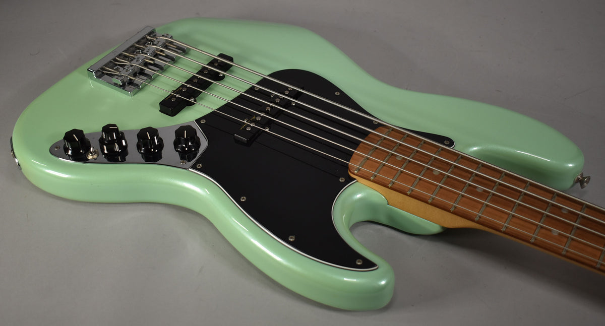 A mint green 5-string bass, angled from the lower bout, showing its black pickguard, chrome bridge, and four black control knobs.