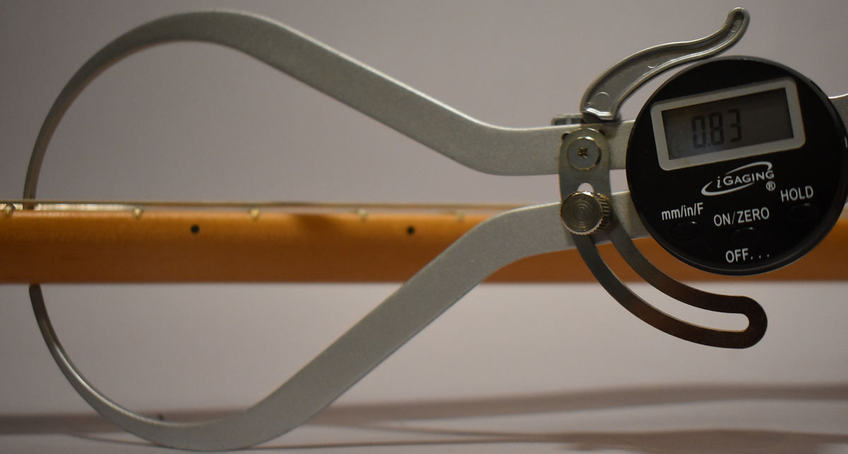 A digital caliper measures the guitar's light brown neck, displaying "0.83" on its black screen, with fret markers visible.
