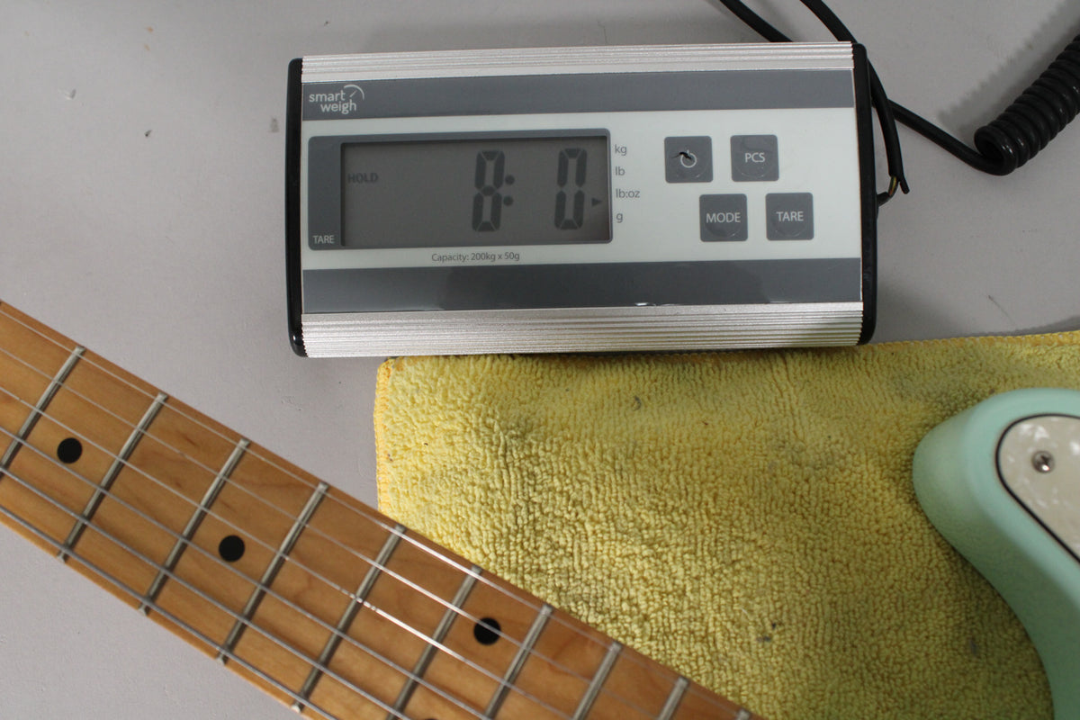 Overhead view of the guitar's maple neck and part of its surf green body, resting on a yellow towel next to a digital scale reading "8:0".