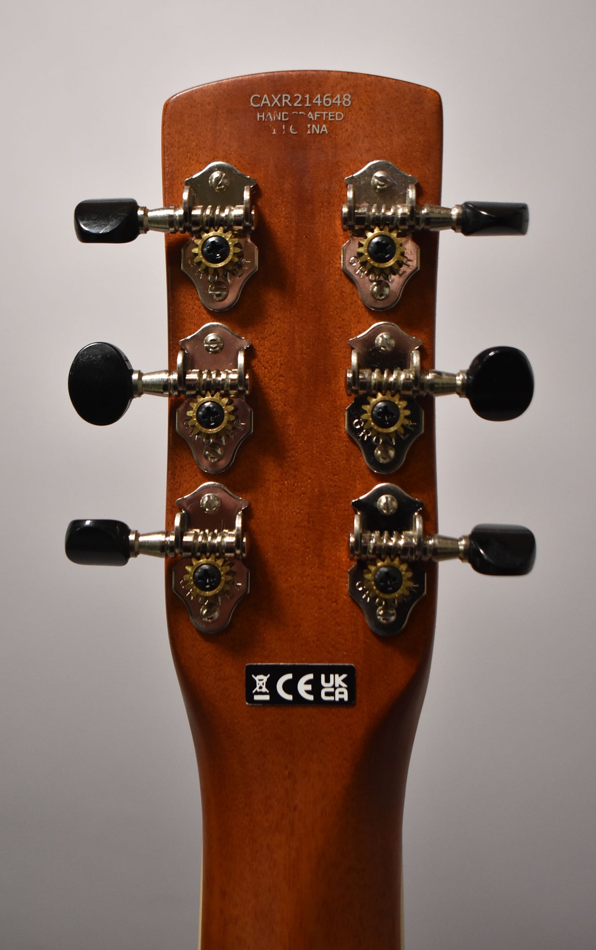 The back of a light brown headstock with six chrome tuners, black buttons, and visible brass gears.