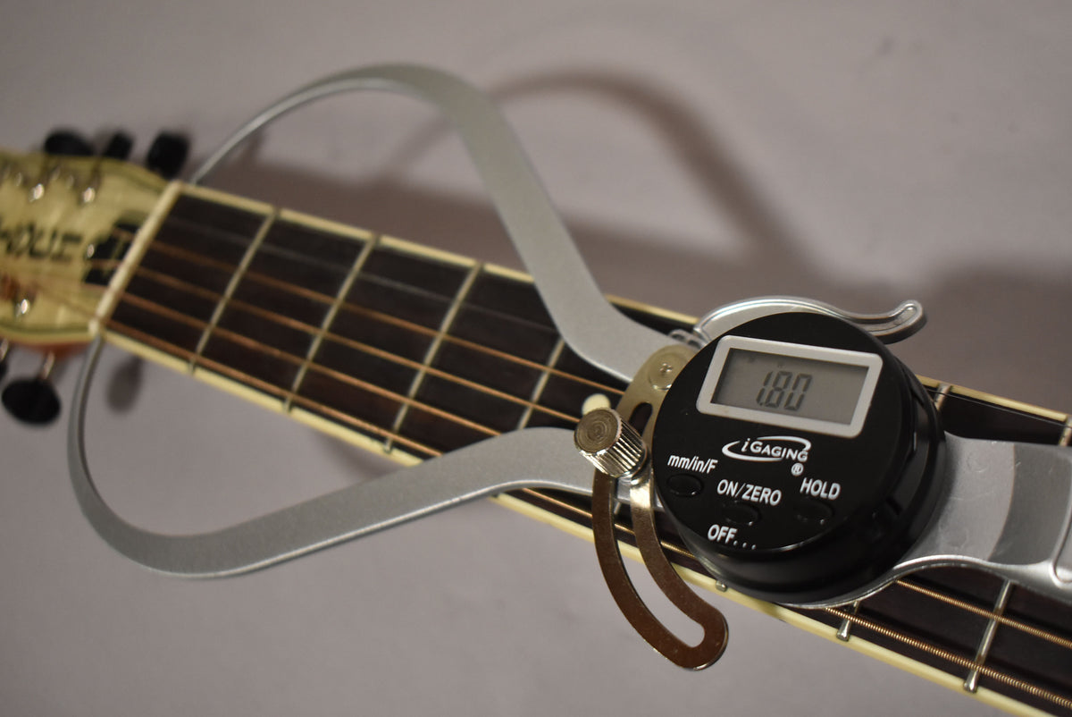 A digital caliper measures the guitar's fretboard, showing "1.00" on its screen, with the headstock and tuning pegs visible.