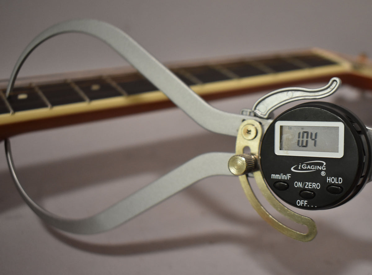 A digital caliper measures the guitar's fretboard, displaying "1.04" on its screen, with the neck angled away.