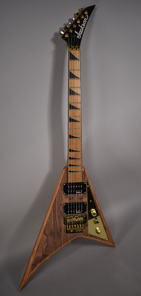 Full-length view of a V-shaped guitar with a natural wood body, gold hardware, black humbuckers, and black sharkfin inlays.