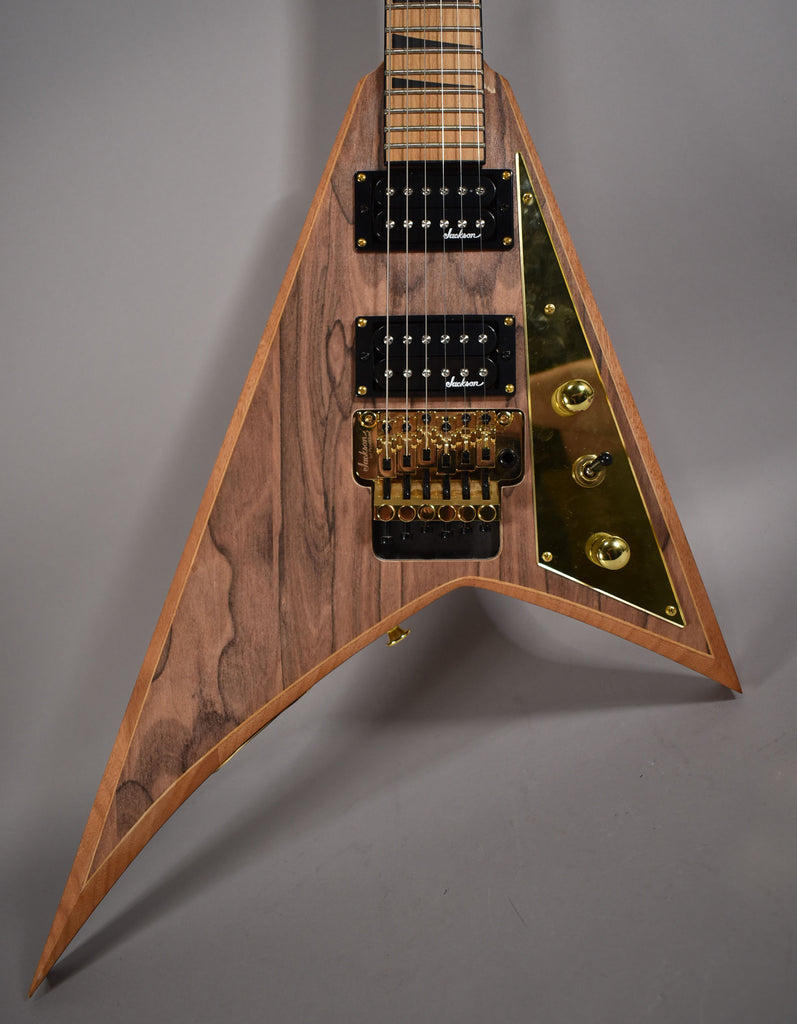 Close-up of the V-shaped body of a natural wood grain guitar with black humbuckers, gold Floyd Rose bridge, and gold control plate.