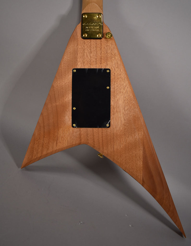 The back of a V-shaped guitar with a light brown wood grain finish, gold neck plate, and black control cavity cover.