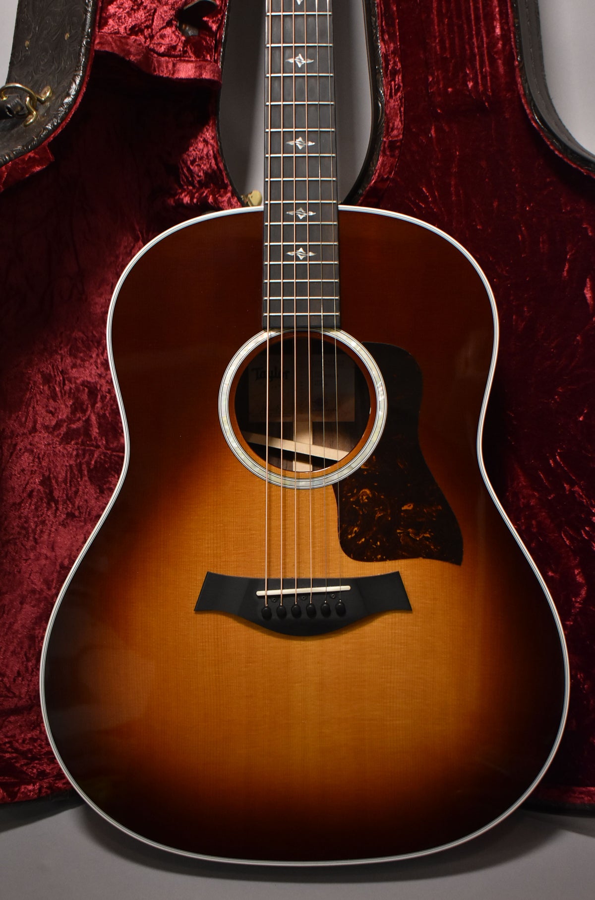 Full front view of a sunburst acoustic guitar with a tortoiseshell pickguard, white binding, and a red velvet case behind it.