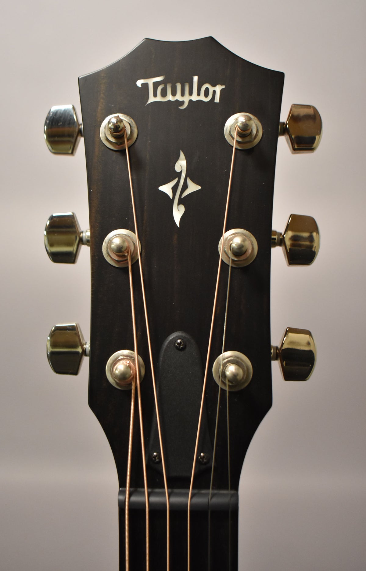 Close-up of the guitar's dark headstock with a white inlaid logo and symbol, six gold tuning pegs, and bronze strings.