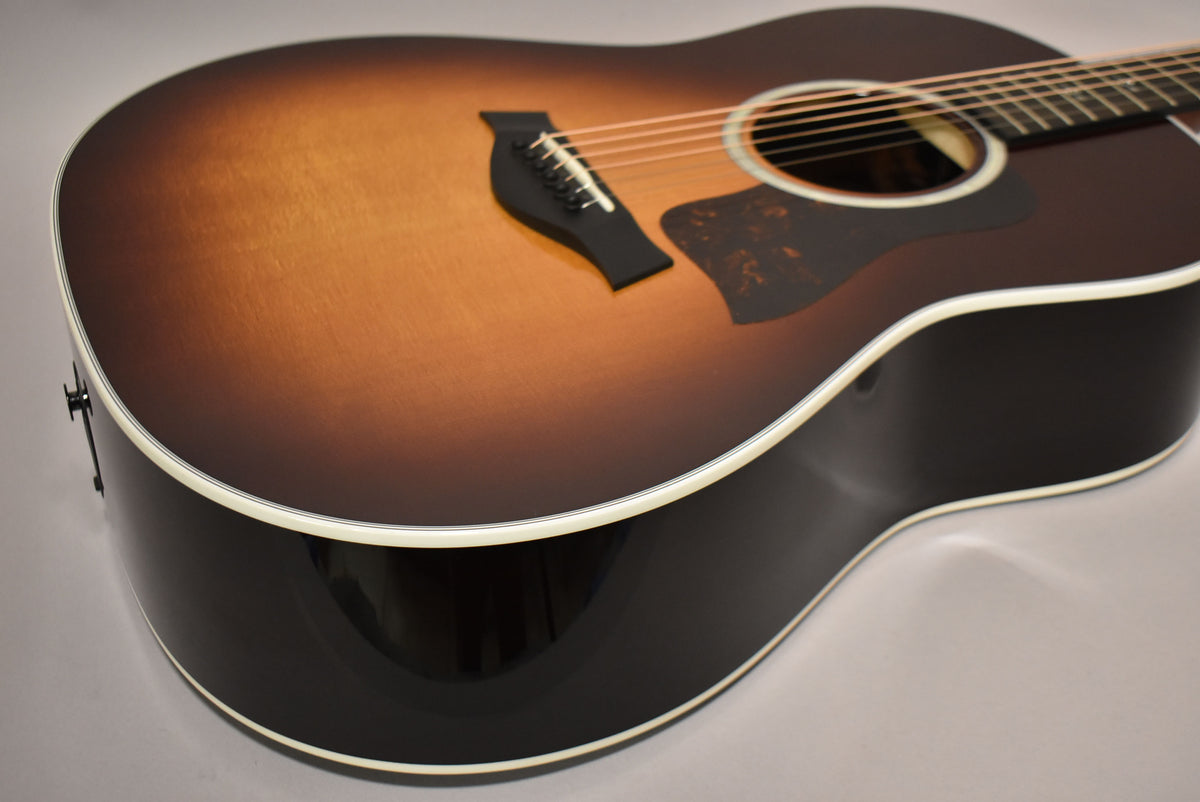 The sunburst acoustic guitar is angled from the lower bout, showing its glossy dark sides and matte top with a black pickguard.