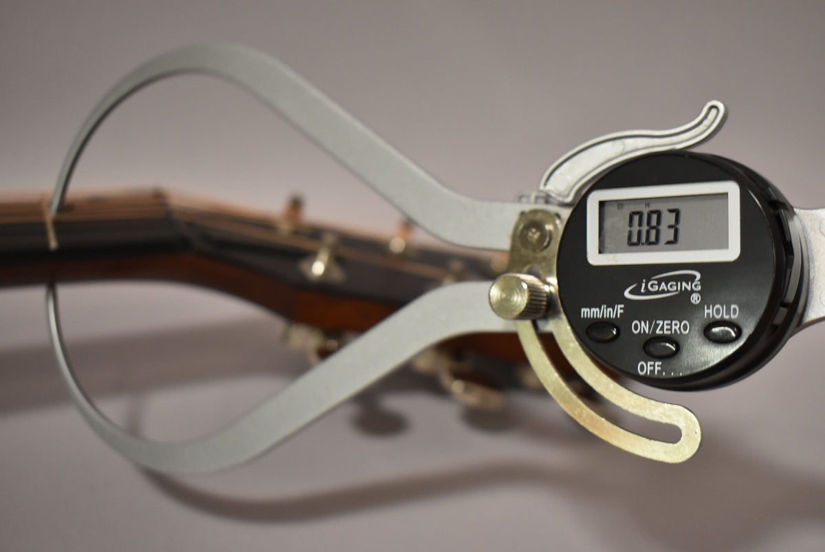 A digital caliper measures the guitar neck, showing 0.83 on its screen, with tuning pegs visible in the background.