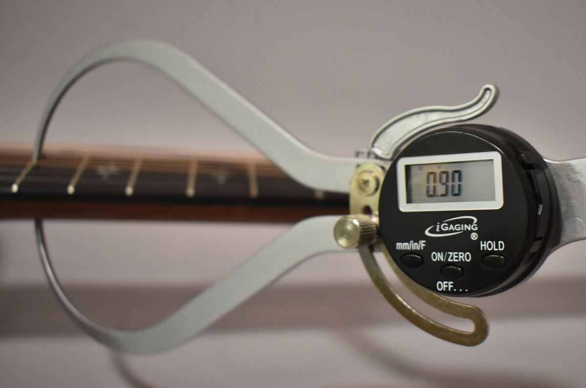 A digital caliper measures the guitar's fretboard, showing "0.90" on its screen, with the frets visible in the background.