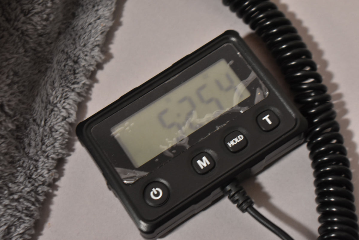 A black digital meter with a display showing "5.254" and buttons for power, M, HOLD, and T, next to a gray fuzzy cloth and a coiled black cable.