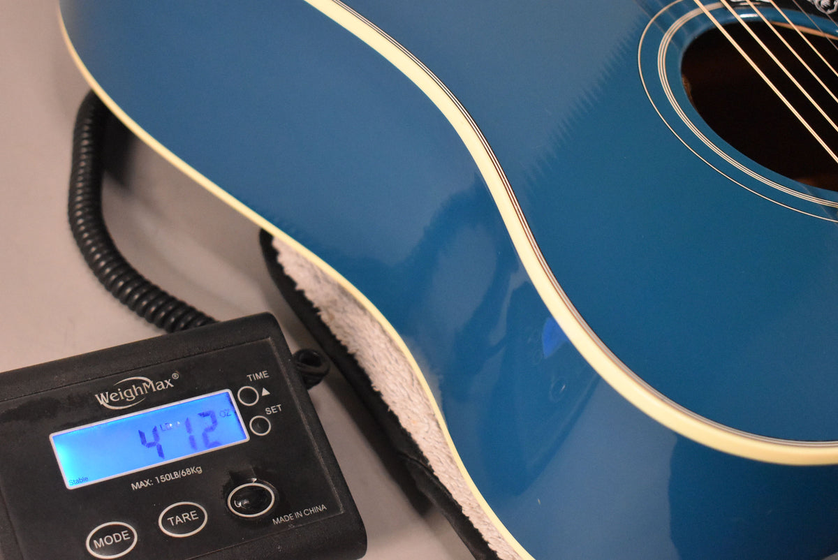 The Big Sky Blue guitar body is shown on a scale displaying "4.72" lbs, with cream binding and a soundhole visible.