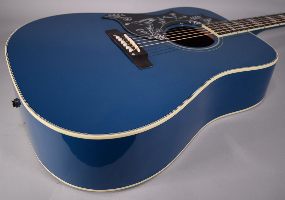 The Big Sky Blue acoustic guitar body, with a white binding and black pickguard with white floral etching, is angled from the lower bout.