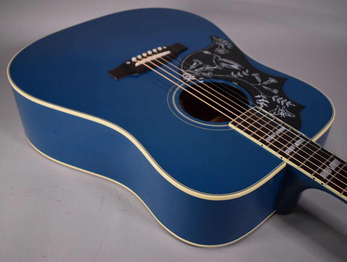 The blue acoustic guitar's body is angled from the upper left, showing the pickguard with white hummingbird and floral etching.