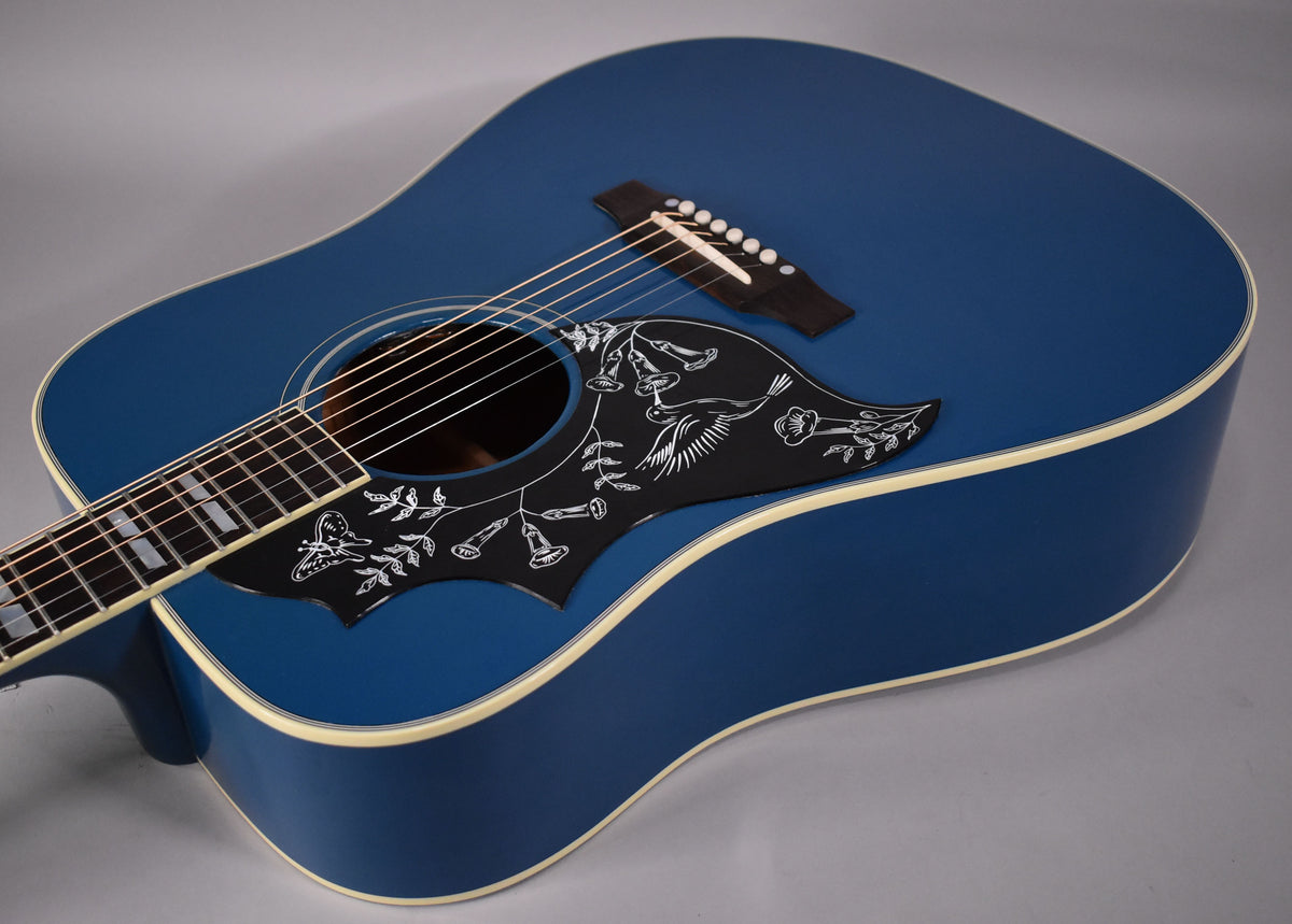 The blue acoustic guitar's body is angled right, showing the black pickguard with white hummingbird and floral etching, and the bridge.