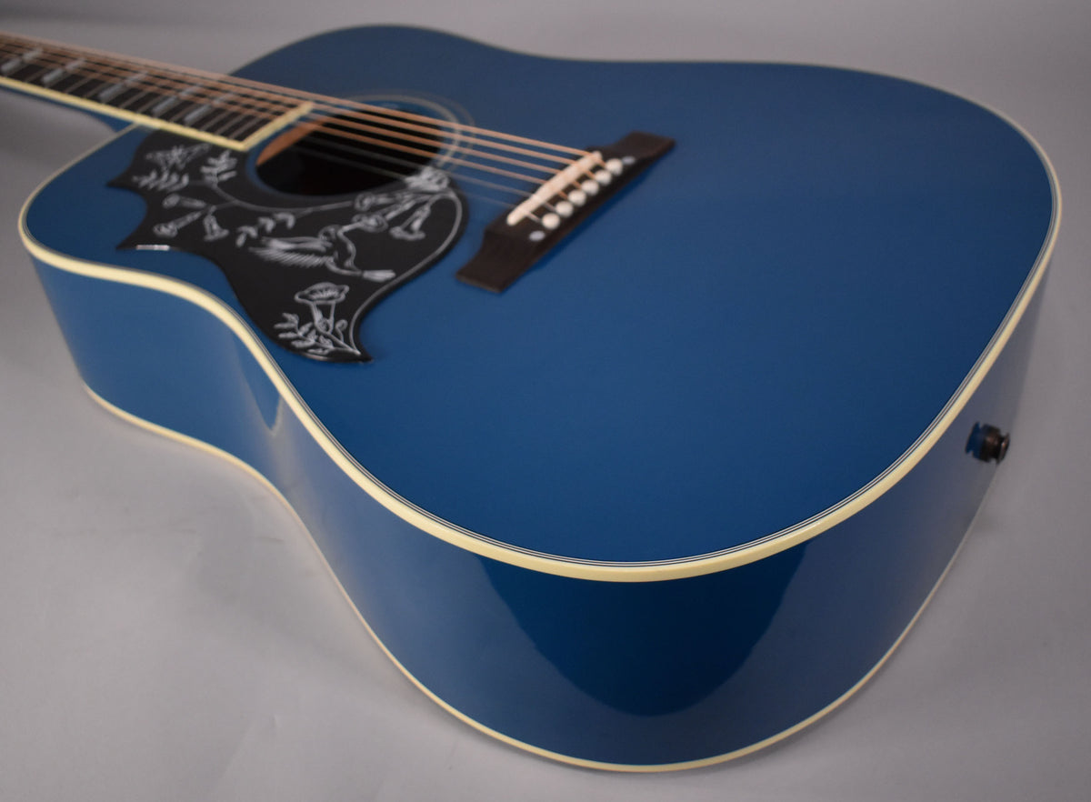 The Big Sky Blue acoustic guitar's body is angled, showing the black pickguard with white etched hummingbird art, and cream binding.