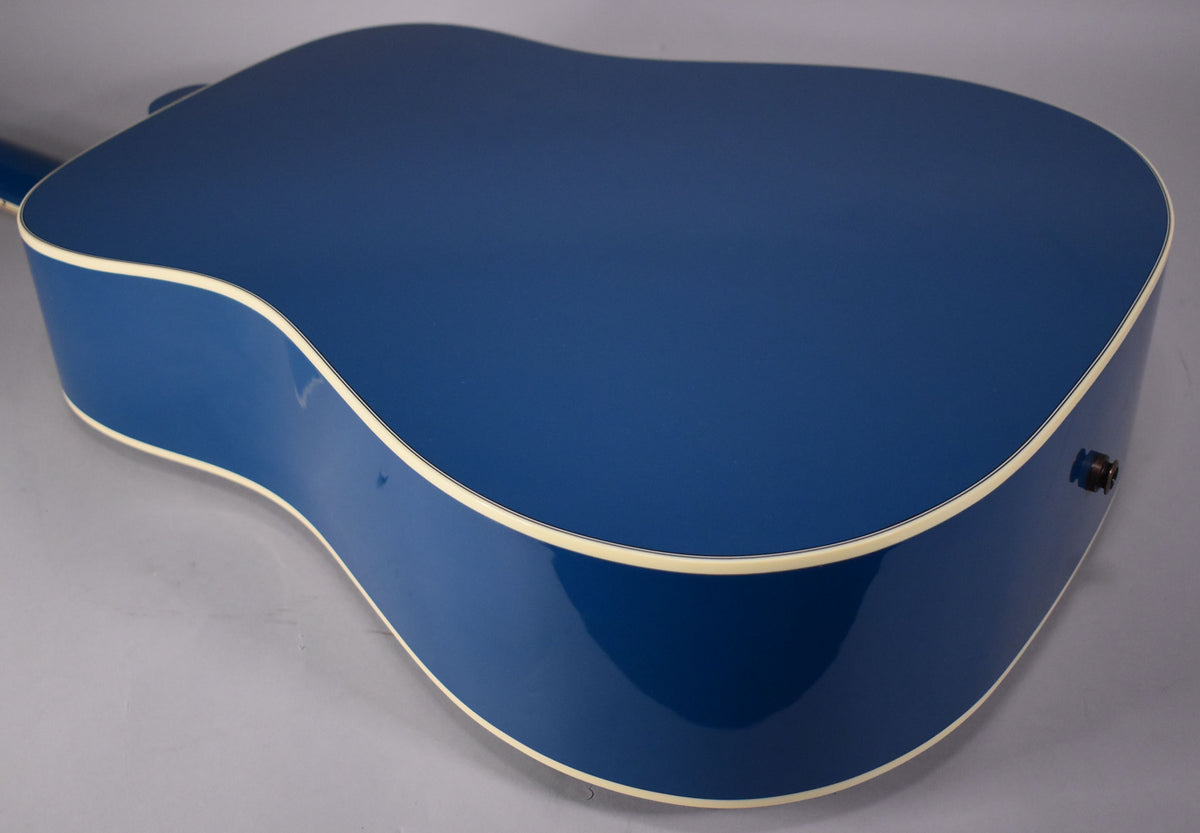 The back and side of a glossy blue acoustic guitar with cream binding, angled from the upper left, showing the endpin jack.