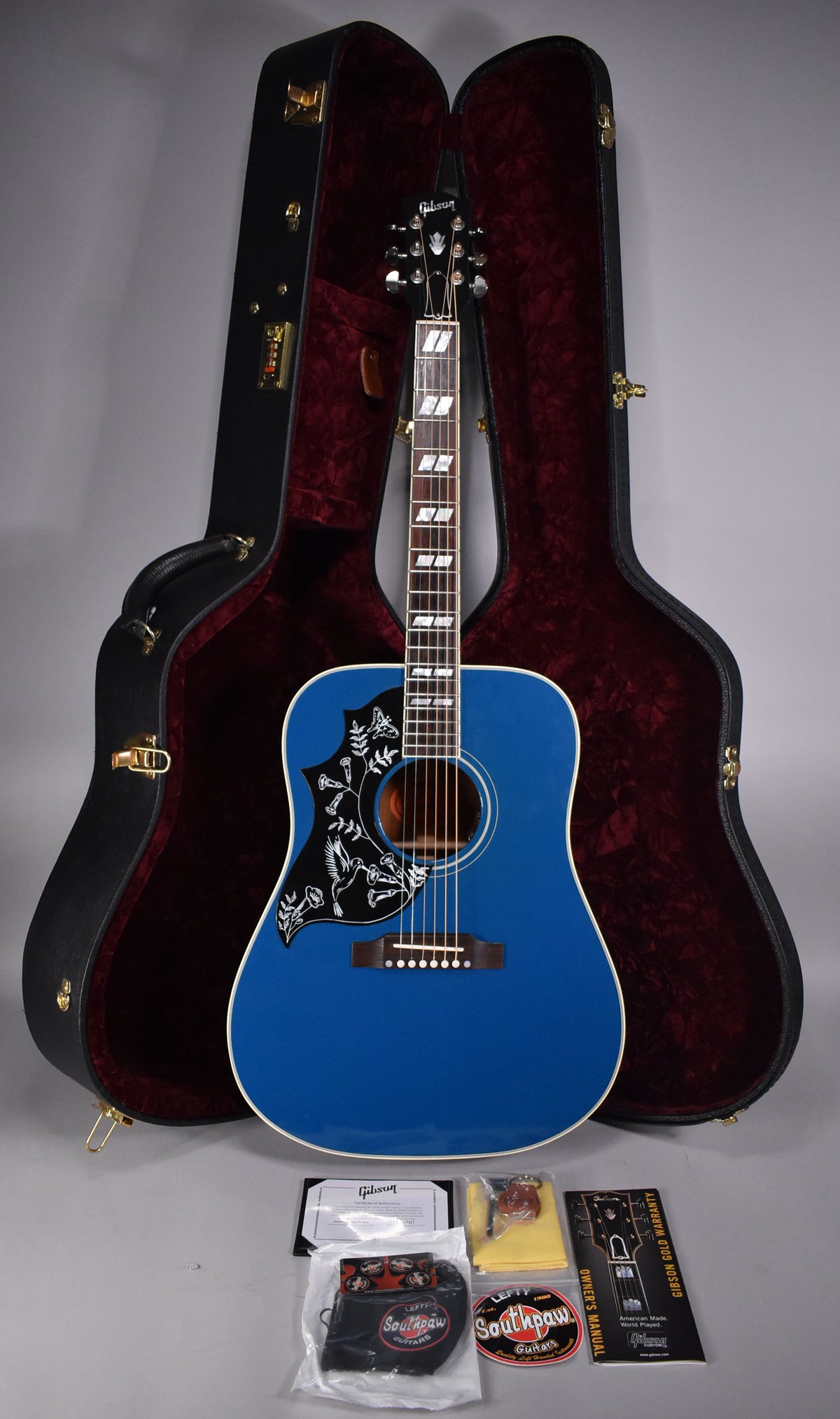 Full view of a Big Sky Blue acoustic guitar with a black pickguard featuring white hummingbirds and flowers, standing in front of its open, red-lined case.