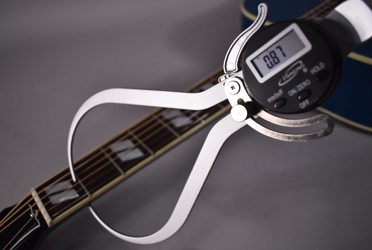 A digital caliper measures the guitar's fretboard, showing 0.87mm, with trapezoid inlays visible on the dark wood.