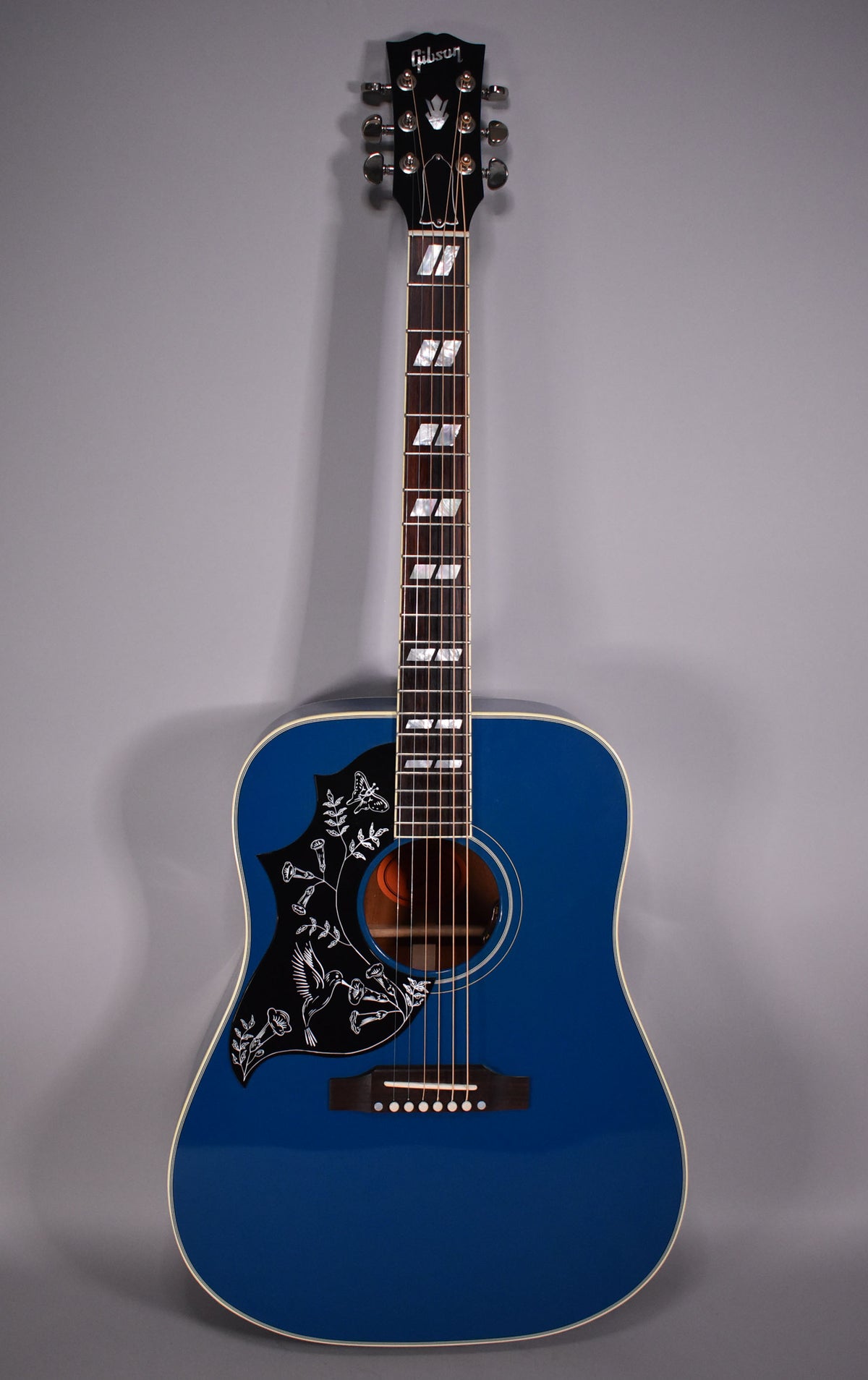 Full front view of a glossy blue acoustic guitar with a black pickguard featuring a white hummingbird and floral design.