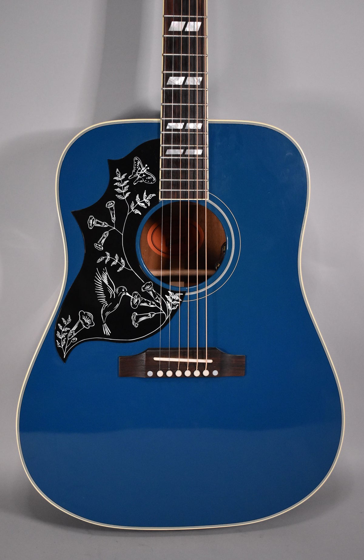 The Big Sky Blue acoustic guitar's body and fretboard are framed, showing a black pickguard with white hummingbird and floral designs.