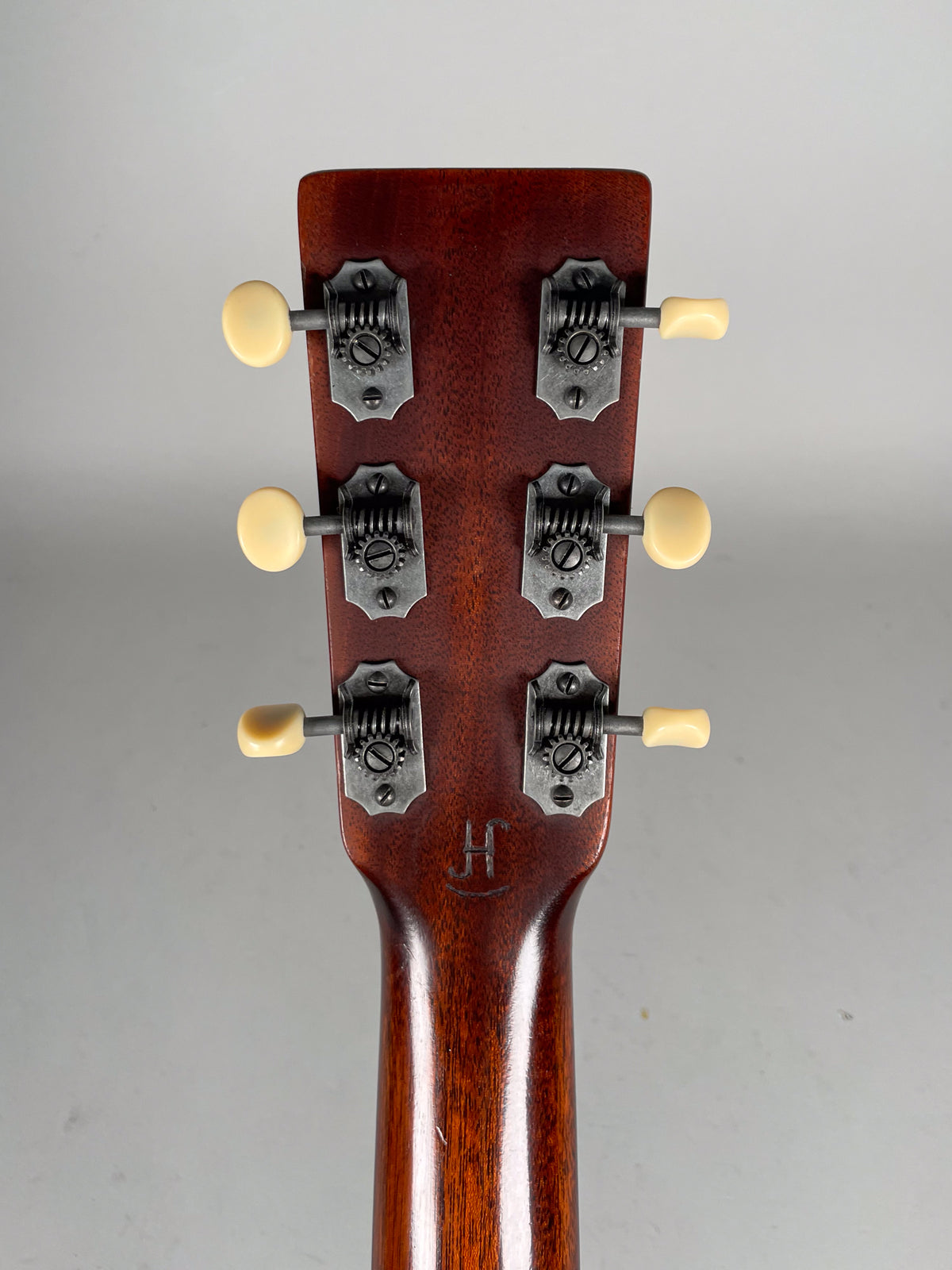 The back of a dark brown mahogany headstock and neck, showing six open-gear tuners with cream buttons and a subtle "H" stamp.