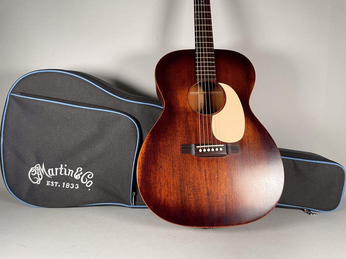 A full-front view of a dark brown acoustic guitar with a cream pickguard, standing next to a black gig bag with blue trim.