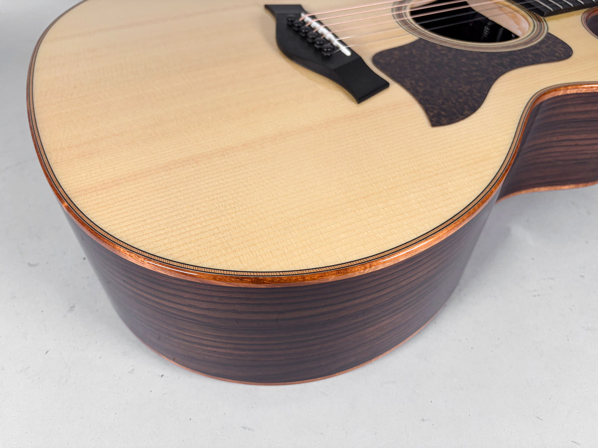 Upper bout of a light-wood acoustic guitar, showing the dark wood sides, black pickguard, and bridge with black pins.