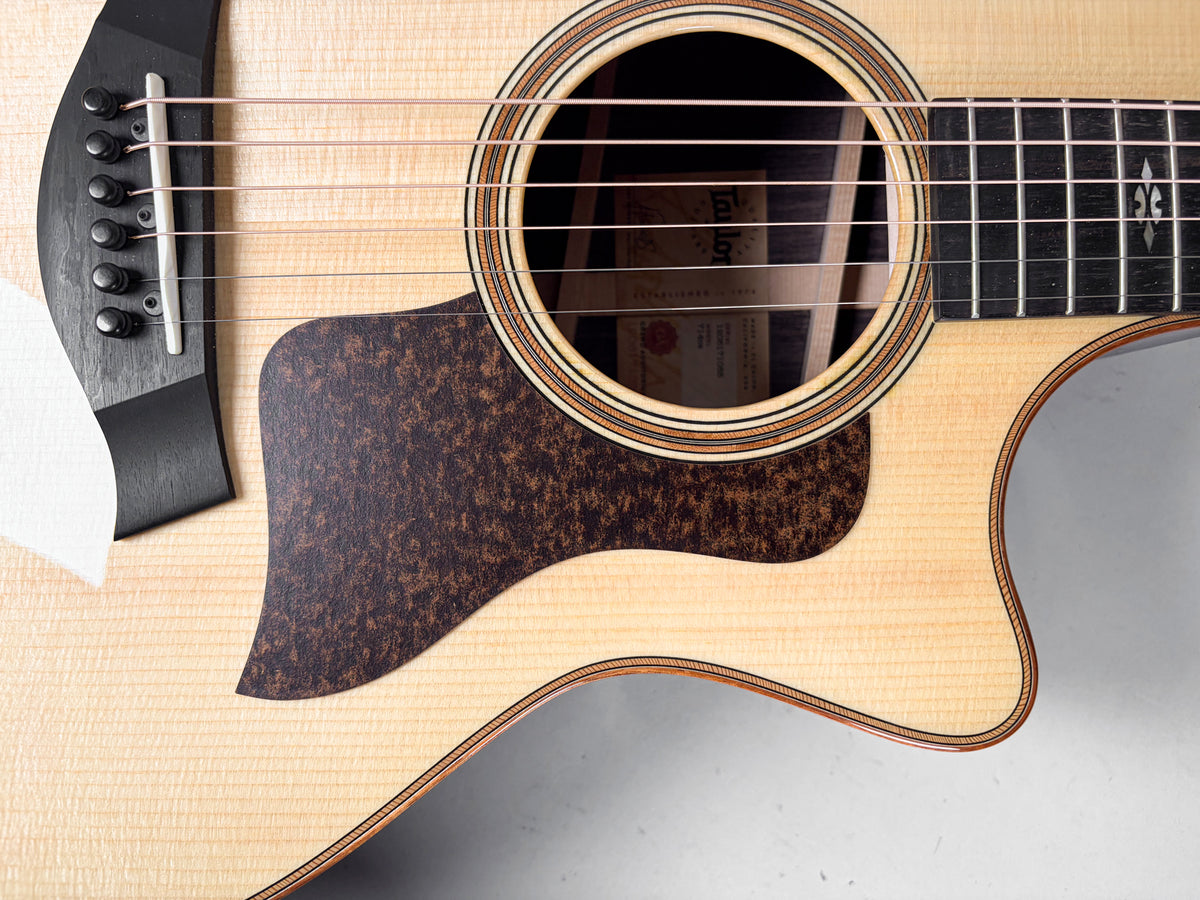 Close-up of a light wood acoustic guitar body, showing the soundhole with a multi-ring rosette, a dark mottled pickguard, and black bridge.